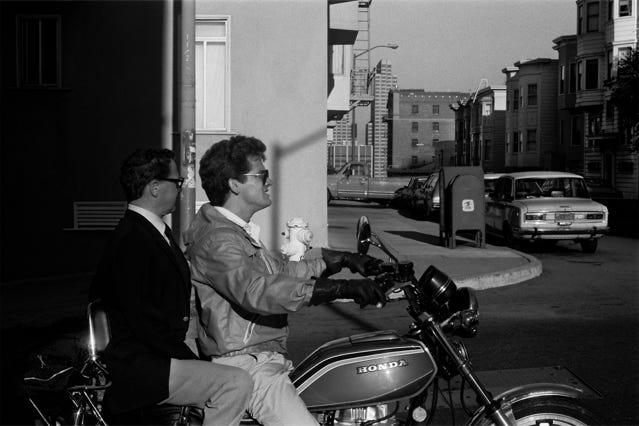 L'image est une photographie en noir et blanc montrant deux hommes sur une moto Honda, dans une rue de San Francisco aux USA en 1980. L'homme au volant porte une veste en cuir et des lunettes de soleil. L'homme à l'arrière porte un costume. Ils sont sur une rue résidentielle avec des voitures garées le long du trottoir et des bâtiments en arrière-plan. La scène est éclairée par un soleil intense, créant des ombres caractéristiques. Le style photographique est réaliste et documentaire, capturant un moment apparemment ordinaire dans la vie urbaine.