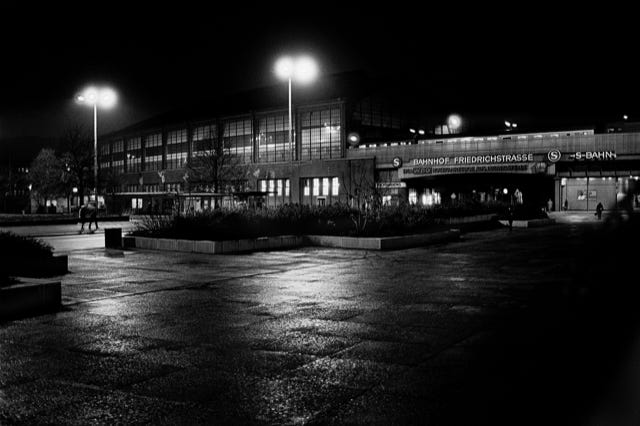 Berlin-Est, RDA, 1980, photographie de Jean pierre Morcrette. Paysage urbain nocturne en noir et blanc devant la gare Friedrichstrasse à Berlin-Est. Des lampadaires éclairent une allée mouillée. Quelques personnes près de l’entrée, avec des plantes en bacs.