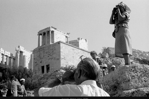 La photo, en noir et blanc, de l’Acropole d’Athènes en 1979, montre, vestiges, colonnes, constructions en pierre et touristes. Un homme photographie le site, tandis qu’une femme sur un rocher prend une photo dans la direction opposée. D’autres visiteurs sont dispersés dans la scène.
