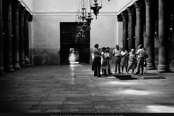 Basilique de la Nativité, Bethléem, Cisjordanie, Territoire occupé, Israël, 1979, photographie de Jean pierre Morcrette.  Cette photo en noir et blanc montre, sur la partie centrale droite, un groupe de personnes rassemblées dans Basilique de la Nativité, où se trouve la grotte de la Nativité, lieu supposé de la naissance de Jésus-Christ, située sous l’édifice. L'intérieur est caractérisé par des colonnes massives et des arches, typiques de l'architecture byzantine. Des lustres ornés pendent du plafond. Un groupe de personnes, touristes ou pèlerins, est rassemblé autour d’un guide à un point central droit, près d’une trappe ouverte. Le sol est pavé de grandes dalles irrégulières, ajoutant un aspect ancien et historique.