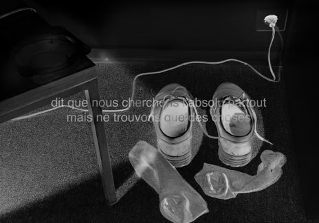 Série métaphysiquée - dit que nous cherchons l'absolu partout mais ne trouvons que des choses, photographie de Jean pierre Morcrette. Photo en négatif noir et blanc montrant des chaussettes et chaussures posées au sol. À gauche on voit l’amorce d’une chaise avec un cendrier vide posé dessus. La scène est traversée par un câble électrique branché à une prise de courant en haut à droite. Deux lignes de texte sont visibles en surimpression au centre de l’image : dit que nous cherchons l'absolu partout mais ne trouvons que des choses