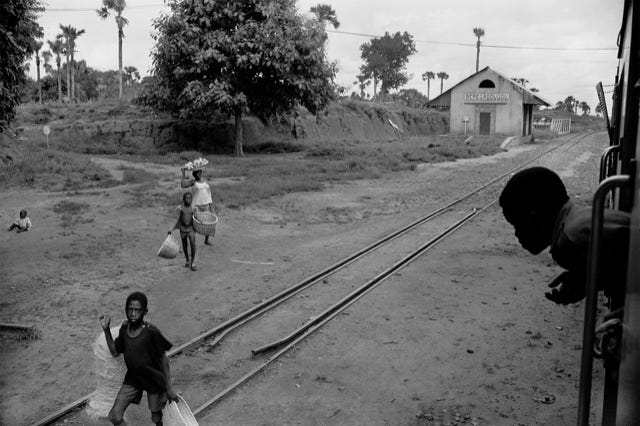 Bérégadougou, Haute-Volta, aujourd'hui Burkina Faso, 1982,  photographie de Jean pierre Morcrette. Cette photo en noir et blanc capture une scène de la vie quotidienne à la gare de Bérégadougou. Au premier plan à droite, on devine un train à l’arrêt d’où sort le buste partiellement visible d’un jeune garçon penché à la fenêtre. Des rails traversent l’image en diagonale du bas gauche au haut droit. Au premier plan à gauche, un jeune garçon marche en portant un objet d’une main et un panier vide de l’autre. Il regarde vers l’objectif du photographe, et semble vouloir proposer à la vente ses marchandises. Au centre de l'image gauche, une femme et un jeune garçon venant d’un chemin marchent côte à côte. L'une porte un plateau chargé de fruits sur la tête, tandis que l'autre tient un panier vide à chaque main. À l’extrême gauche, au centre près du bord de l’image, un bébé est assis au sol, seul. Le paysage est rural, avec des arbres et des palmiers dispersés. Le sol est poussiéreux, typique des zones sèches ou semi-arides.