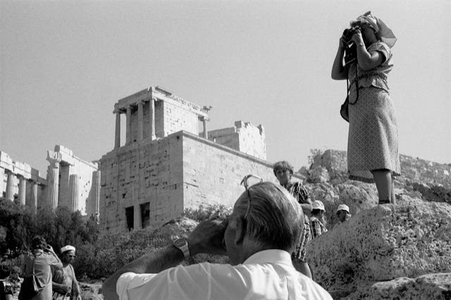 Acropole, Athènes, Grèce, 1979, photographie de Jean pierre Morcrette. La photo, en noir et blanc, montre, vestiges, colonnes, constructions en pierre et touristes. Un homme photographie le site, tandis qu’une femme sur un rocher prend une photo dans la direction opposée. D’autres visiteurs sont dispersés dans la scène.