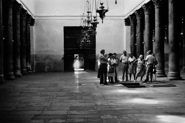 Basilique de la Nativité, Bethléem, Cisjordanie, Territoire occupé, Israël, 1979, photographie de Jean pierre Morcrette.  Cette photo en noir et blanc montre, sur la partie centrale droite, un groupe de personnes rassemblées dans Basilique de la Nativité, où se trouve la grotte de la Nativité, lieu supposé de la naissance de Jésus-Christ, située sous l’édifice. L'intérieur est caractérisé par des colonnes massives et des arches, typiques de l'architecture byzantine. Des lustres ornés pendent du plafond. Un groupe de personnes, touristes ou pèlerins, est rassemblé autour d’un guide à un point central droit, près d’une trappe ouverte. Le sol est pavé de grandes dalles irrégulières, ajoutant un aspect ancien et historique.