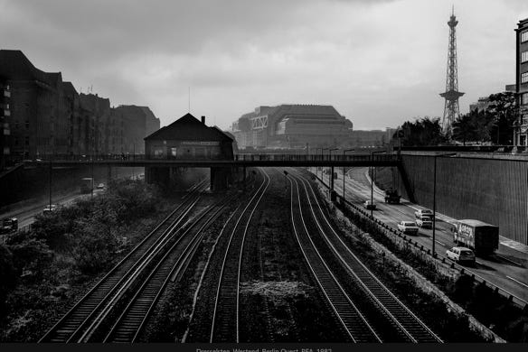 Berlin-Ouest, 1982 : vue urbaine en noir et blanc, avec voies ferrées et routes convergeant vers l’horizon, un pont piétonnier et des bâtiments modernes, sous un ciel brumeux.